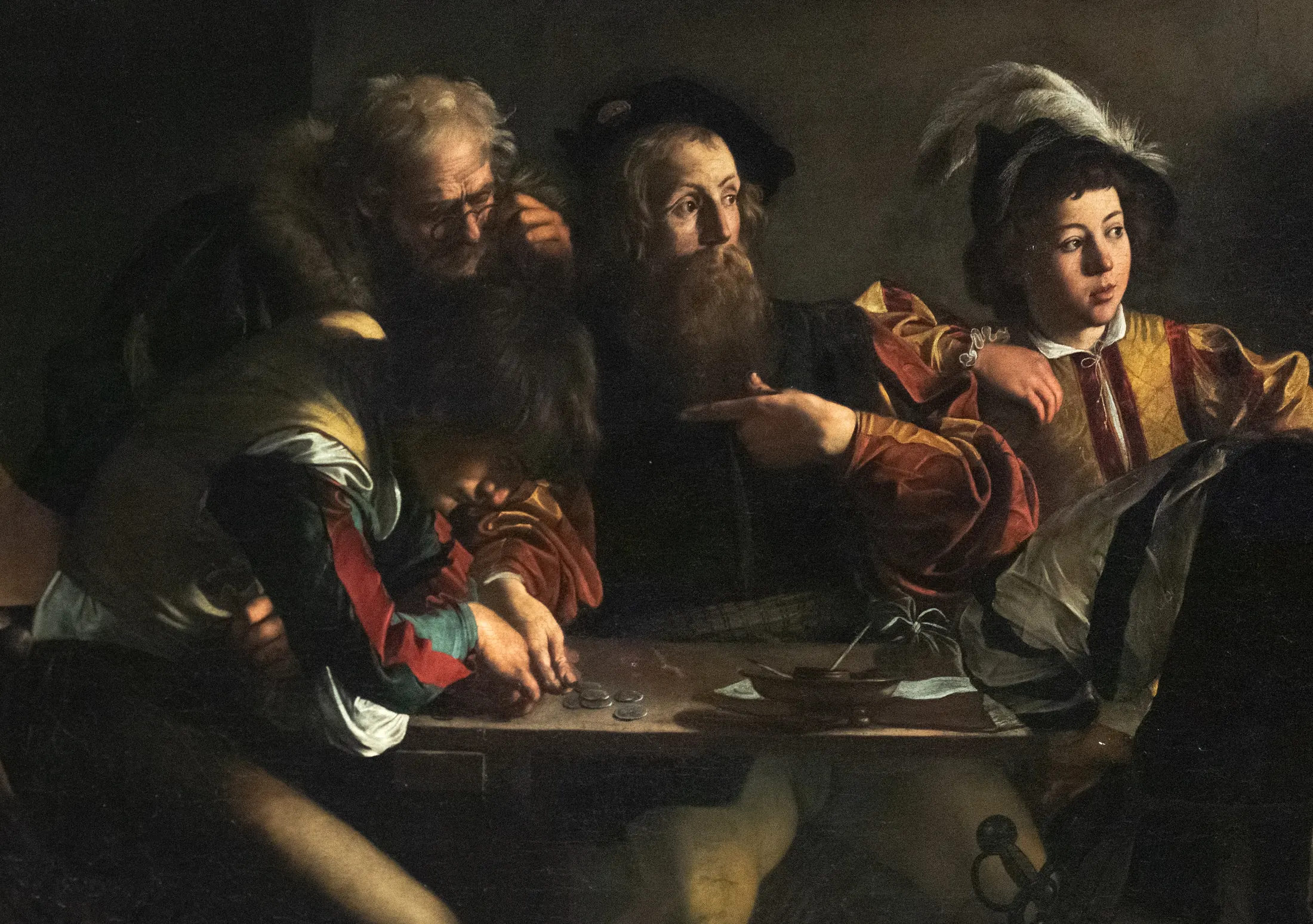 Detail of The Calling of Saint Matthew: the group at the table, with the bearded man in the middle turning toward the light and pointing a hand that could mean himself or the man beside him, an old man in spectacles leaning in, and at the left end a young man bent low over the coins on the table without looking up.