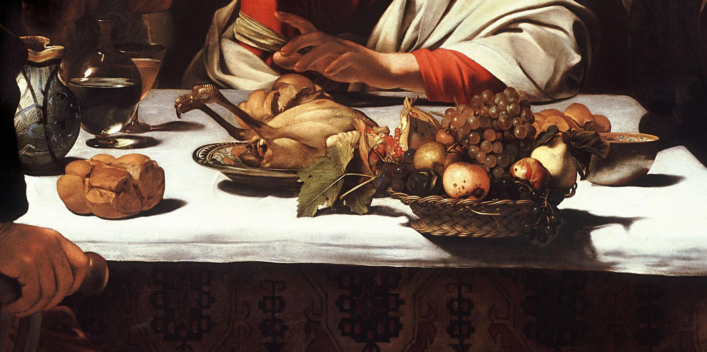 Detail of the Supper at Emmaus: the roast fowl, the split pomegranate, and the basket of bruised and wormholed fruit pushed out over the front edge of the table. Every object throws its own hard shadow across the white cloth, which is what fixes them in the room.