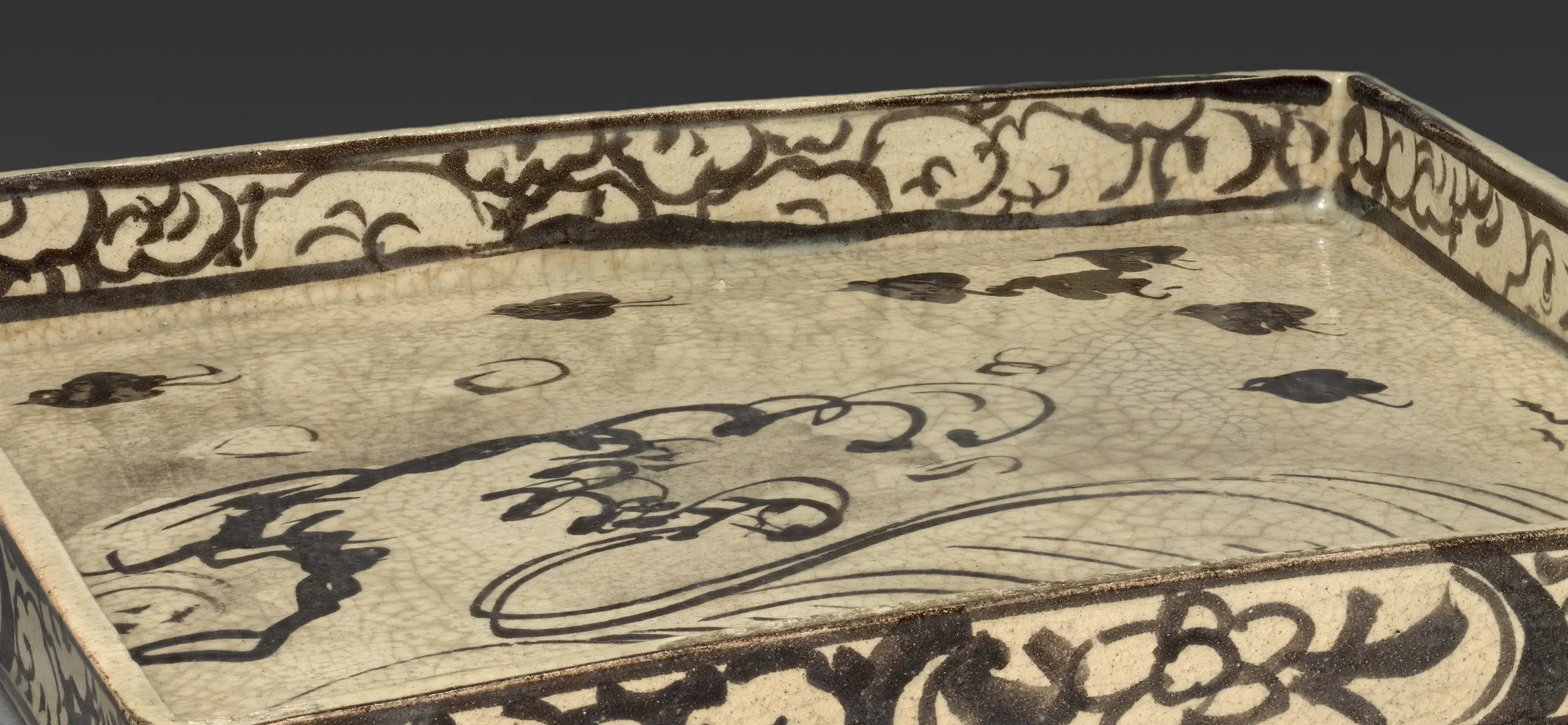 Detail of a square serving dish from a set for tea cakes: plovers over a wave, put down by Kōrin in iron oxide on the unfired clay and then sealed under a clear glaze. Cleveland calls the drawing freely done, and on raw clay there is nothing else it could be. Absorbent, unfired body, no wiping back, no second pass. Every line is the first line.