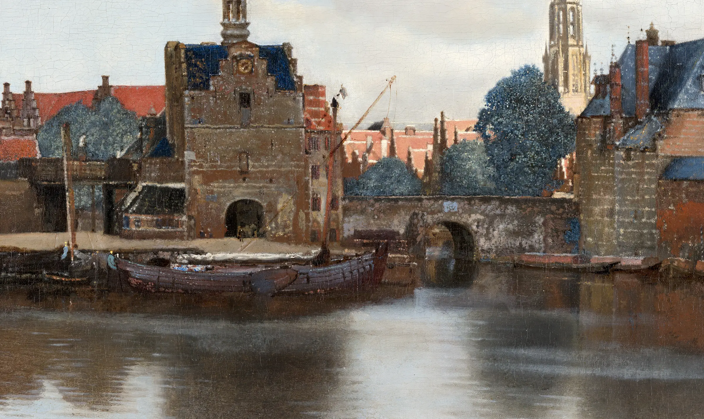 Detail of View of Delft along the water: the gate and the near quay are in shadow and the roofs behind them are in sun, and the line between the two runs right through the middle of the picture. The mortar in the wall, the roof tiles and the hull of the near boat are laid on thick enough to catch a raking light, and the reflection below is its own flat shape rather than a copy of the boat.